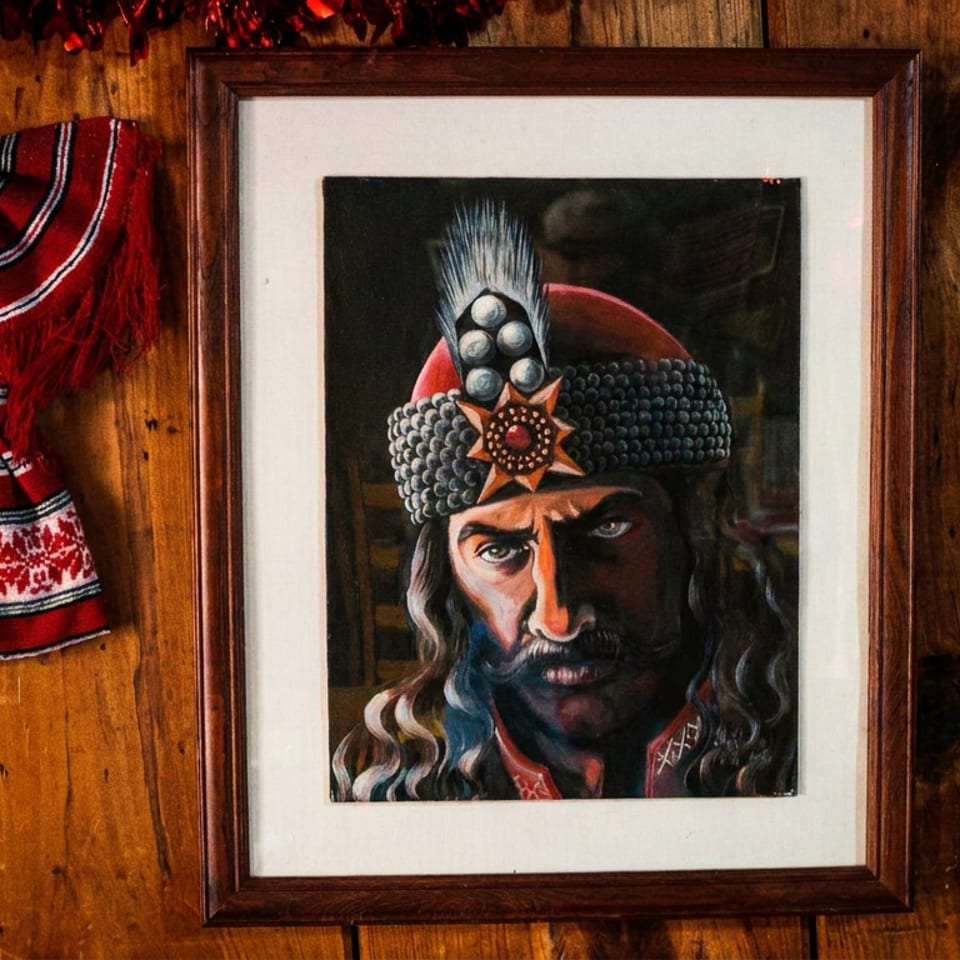 Framed portrait of a mustached man in an ornate traditional hat beside a red embroidered textile on a rustic wooden wall.