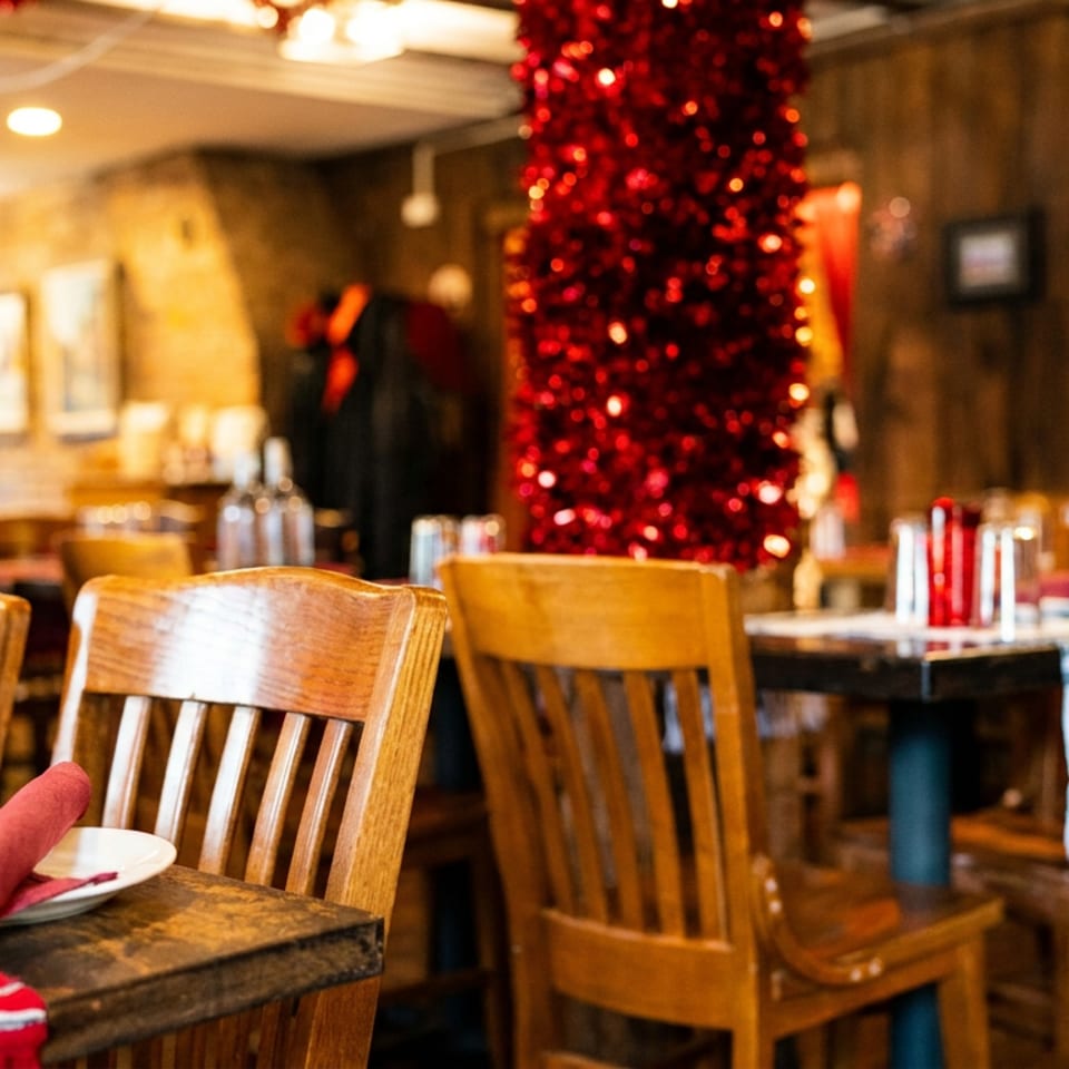 Warmly lit restaurant dining room with wooden tables, red napkins, and festive red garlands wrapped around columns.