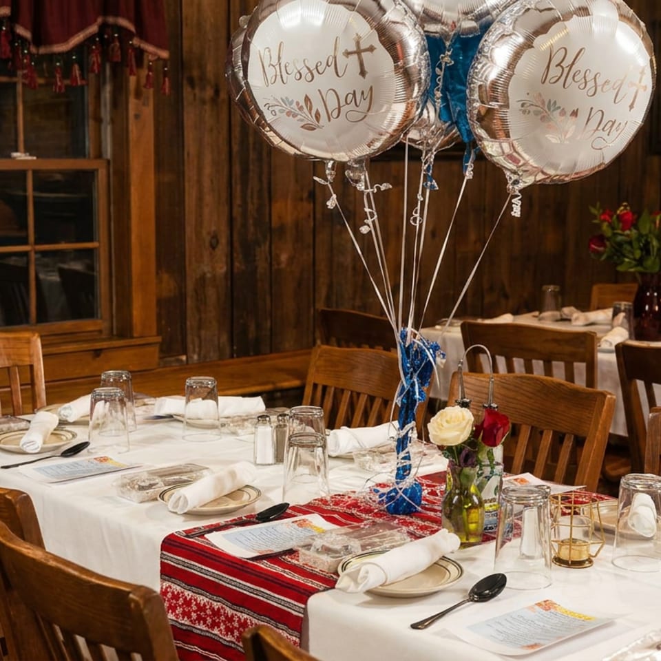 Rustic restaurant dining room with white tablecloths, red runners, floral centerpieces, and celebratory balloons reading “Blessed Day.”