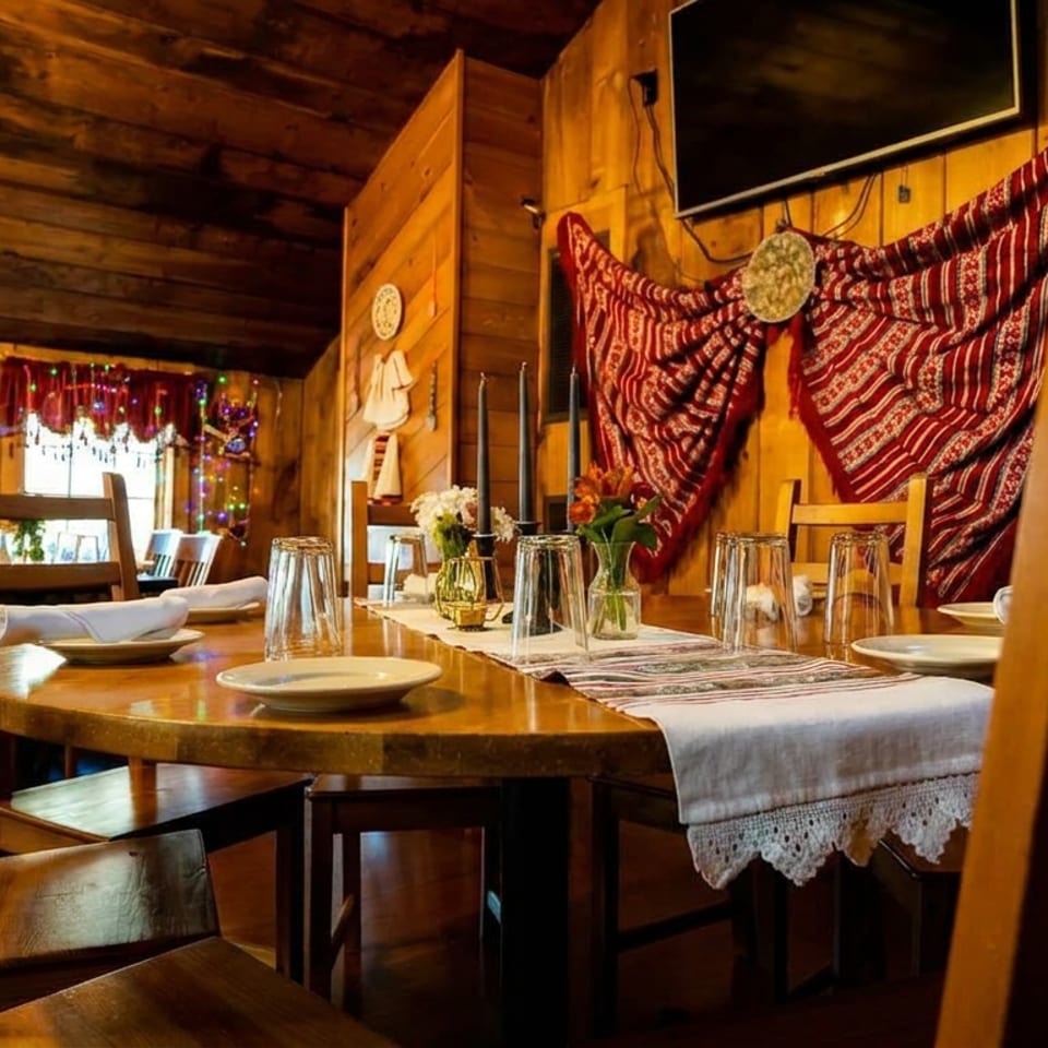 Warm rustic restaurant dining room with wooden tables, set plates, floral centerpieces, string lights, and decorative woven wall hanging.