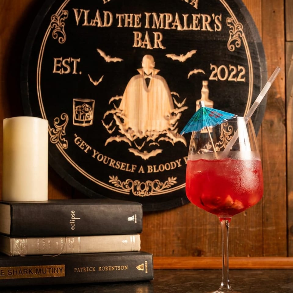 Vivid red cocktail with a blue umbrella beside stacked books and a potted plant, beneath Vlad the Impaler’s Bar sign.