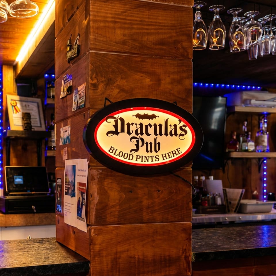 Warmly lit Dracula’s Pub bar interior with hanging wine glasses, liquor shelves, blue accent lights, and a prominent illuminated sign.